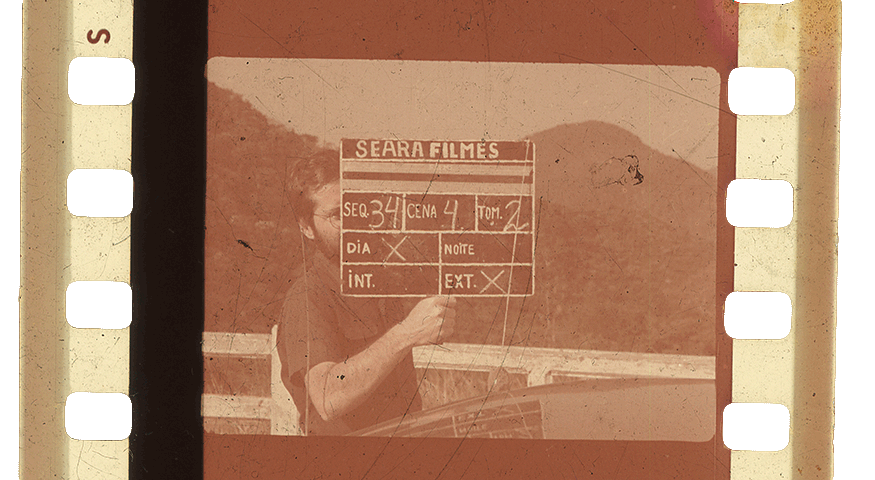 Recorte de negativo de imagem no qual o cineasta Ramon Alvarado, em set de filmagem, está segurando uma claquete na qual lê-se "Seara Filmes" e informações sobre a cena filmada.