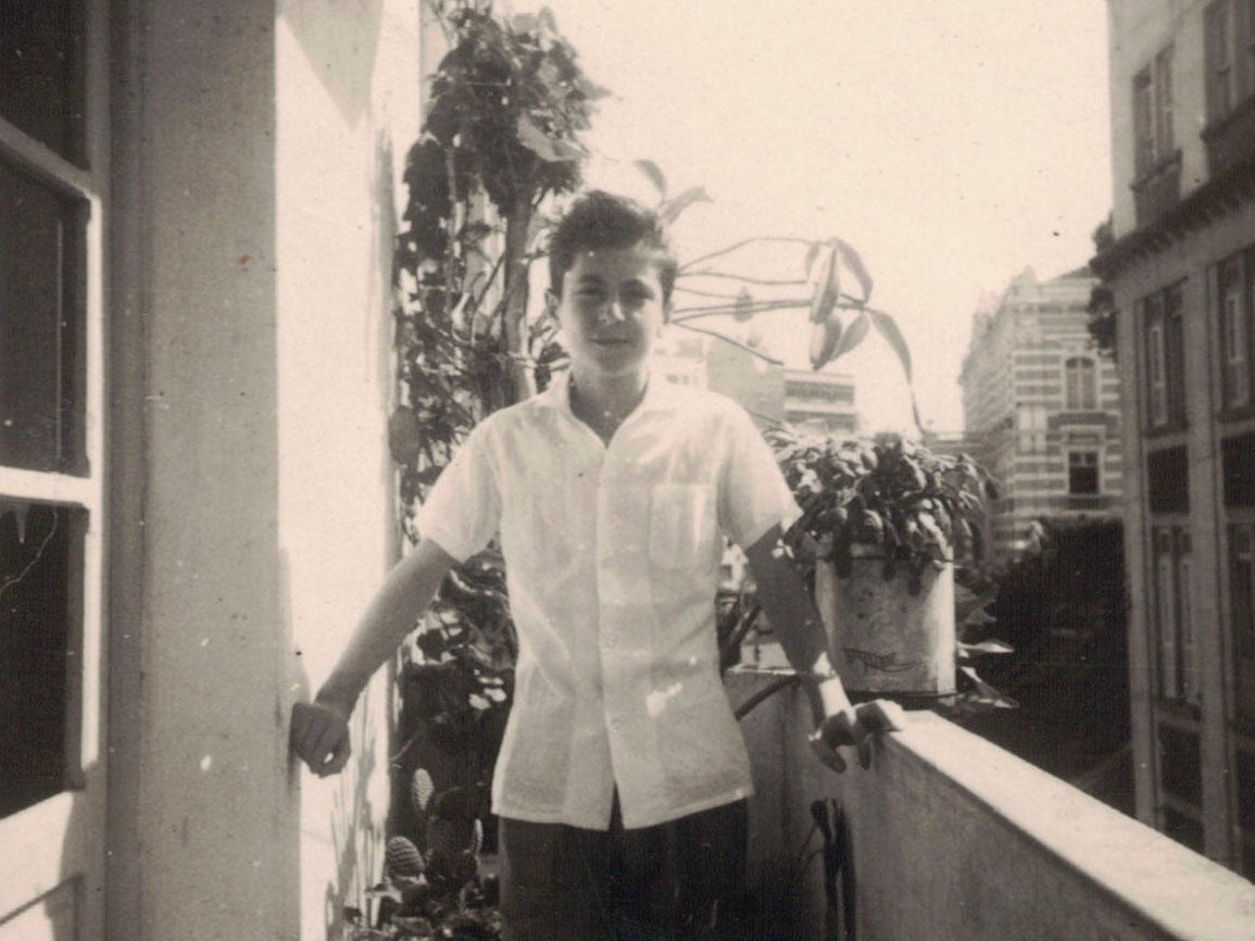 Ramon Alvarado quando criança, vestindo camisa social de mangas curtas, na varanda da casa de seus pais em Vitória (ES), com plantas ao seu lado. Prédios ao fundo, entre eles o Palácio Anchieta.