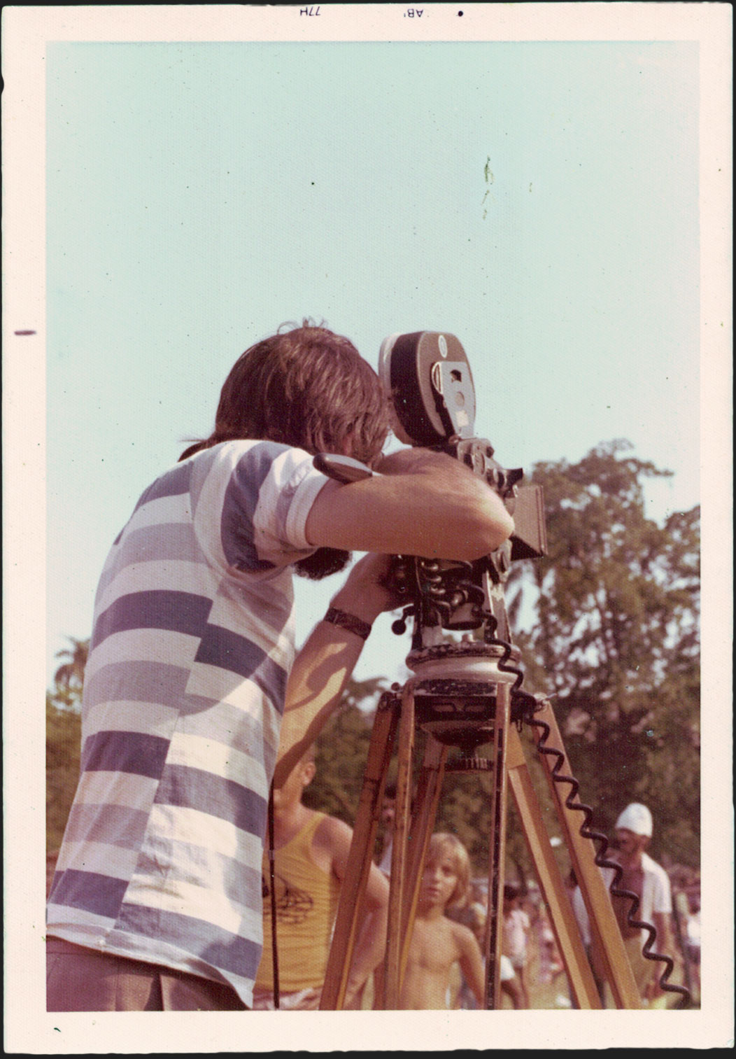 Ramon Alvarado, de costas, opera uma câmera cinematográfica que está sob um tripé. Ao fundo, uma criança olha para o cineasta e seu equipamento e há outros dois homens à sua volta. Ambiente externo, com céu e árvores ao fundo. Fotógrafo(a) não identificado(a). 